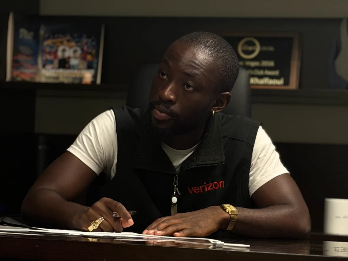 A man wearing a black Verizon vest over a white t-shirt sits at a desk holding a pen and looking thoughtfully to the side.