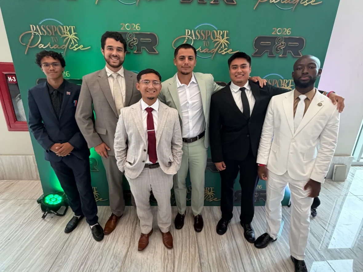 Six sharply dressed men in formal suits pose together in front of a green "Passport to Paradise 2026" event backdrop.