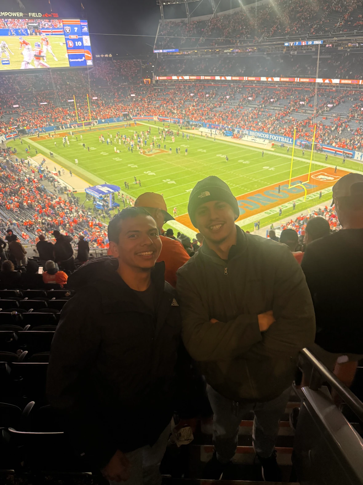 Two smiling young men standing in the stands of a crowded football stadium during an evening game.