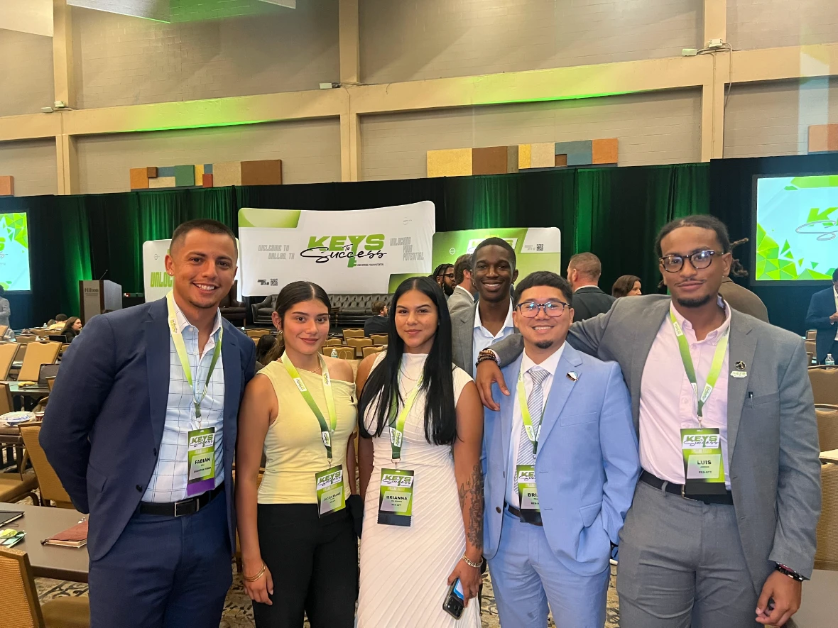Six smiling colleagues in business attire wearing conference name badges pose together in a large convention hall featuring "Keys to Success" branding.
