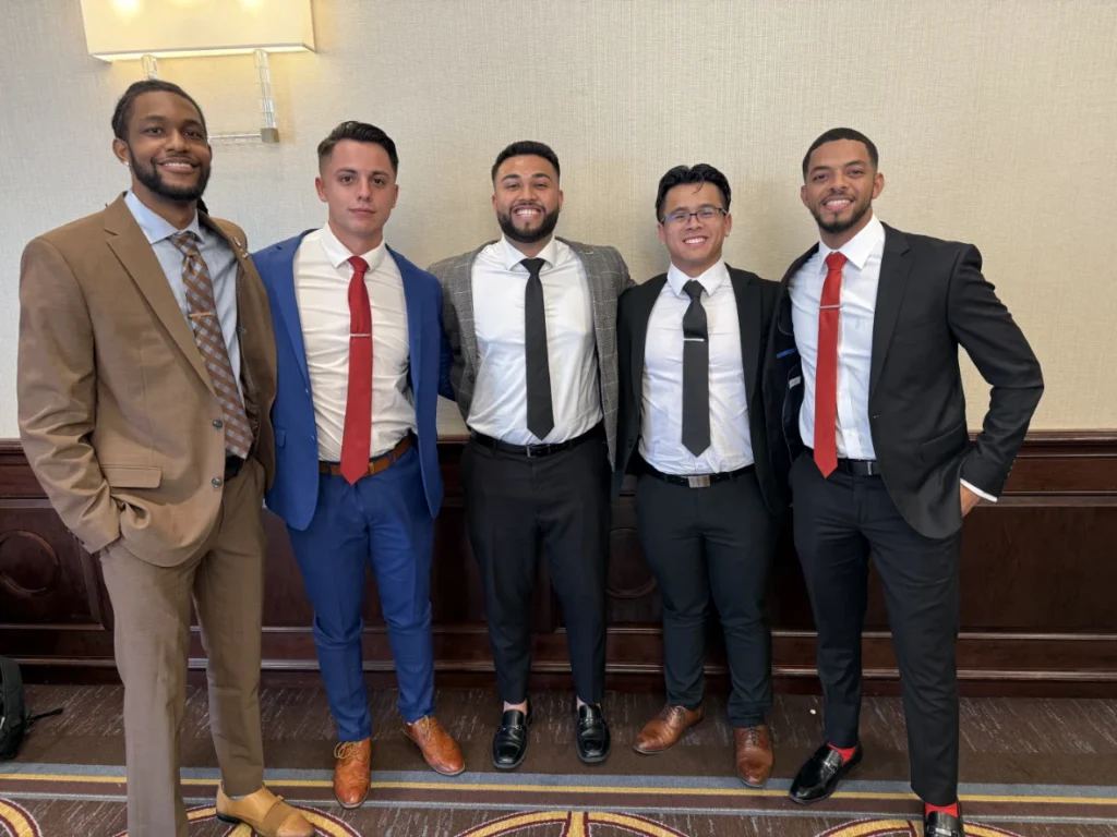 Five men dressed in suits and ties smiling together while posing indoors against a wall with wooden wainscoting.