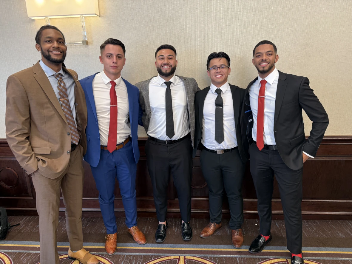 Five men dressed in suits and ties smiling together while posing indoors against a wall with wooden wainscoting.