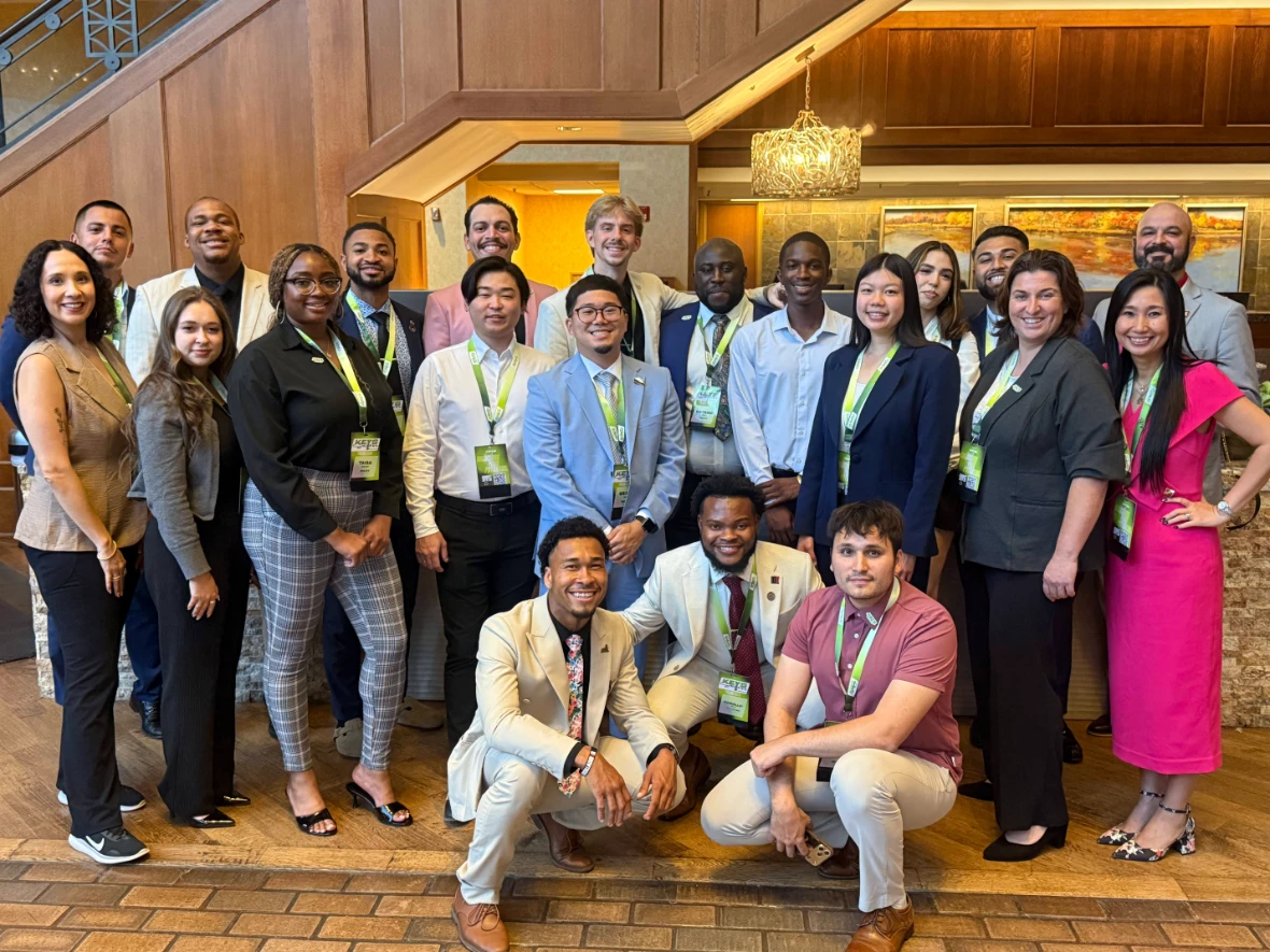 A large group of diverse young professionals in business attire and event lanyards smiling together for a group photo in a lobby.