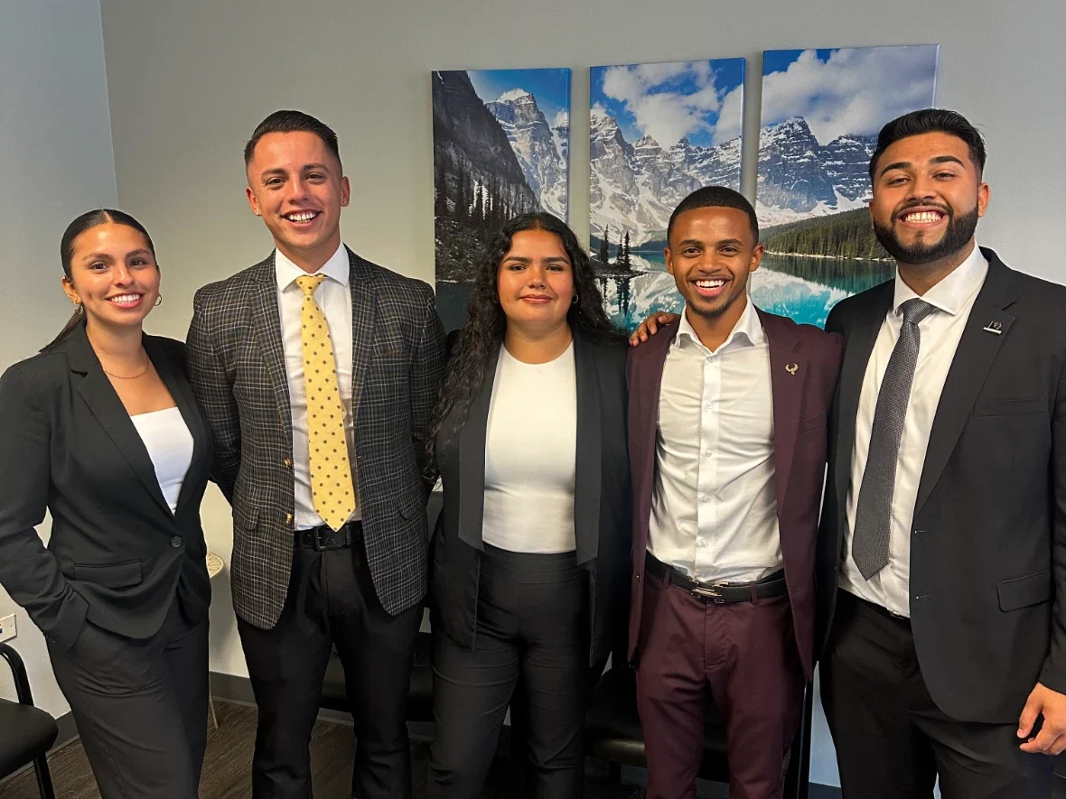 Five smiling colleagues in professional attire pose for a group photo indoors.