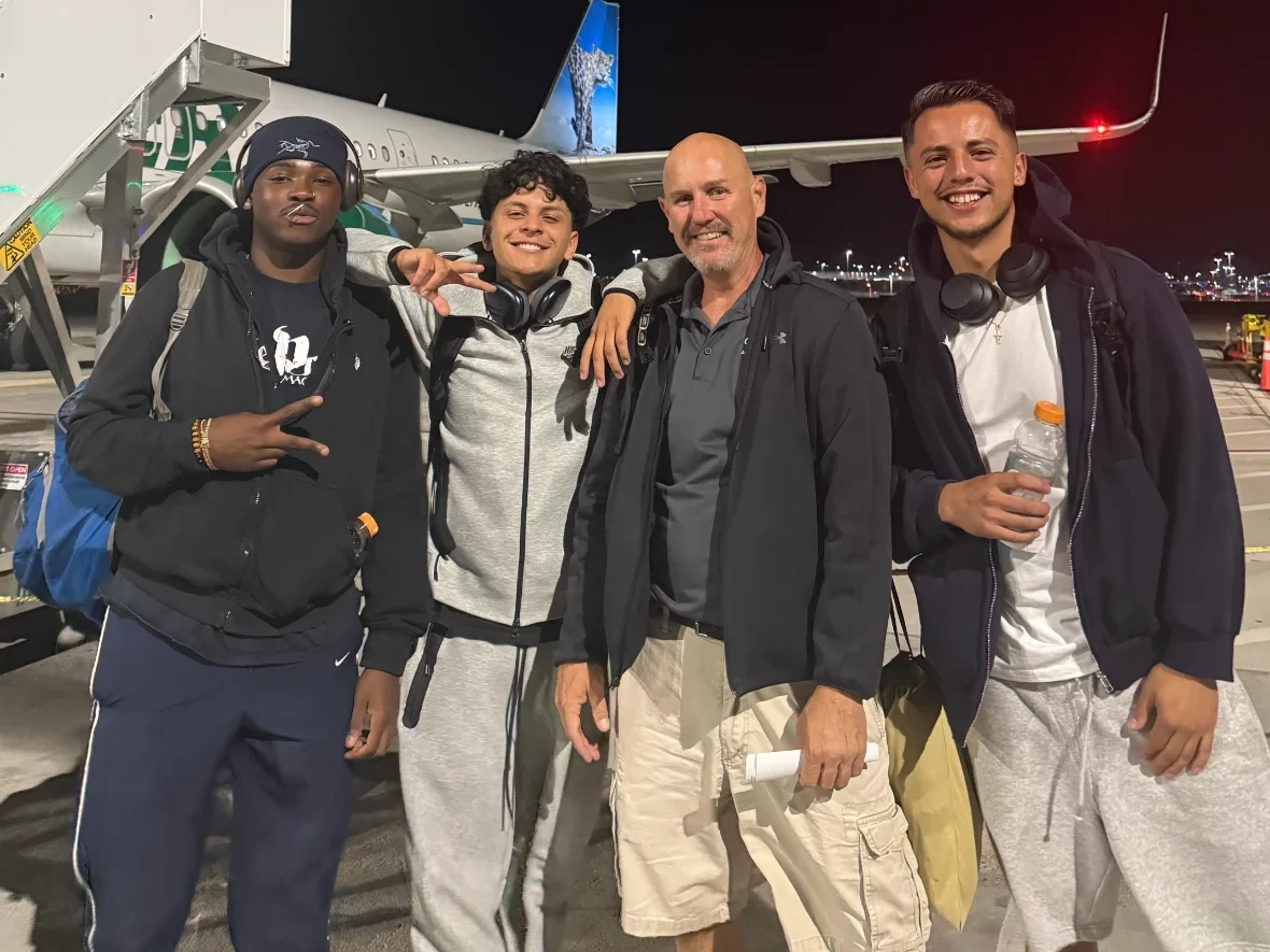 Four men smiling together on an airport tarmac at night, standing in front of an airplane boarding stairway with an aircraft in the background.